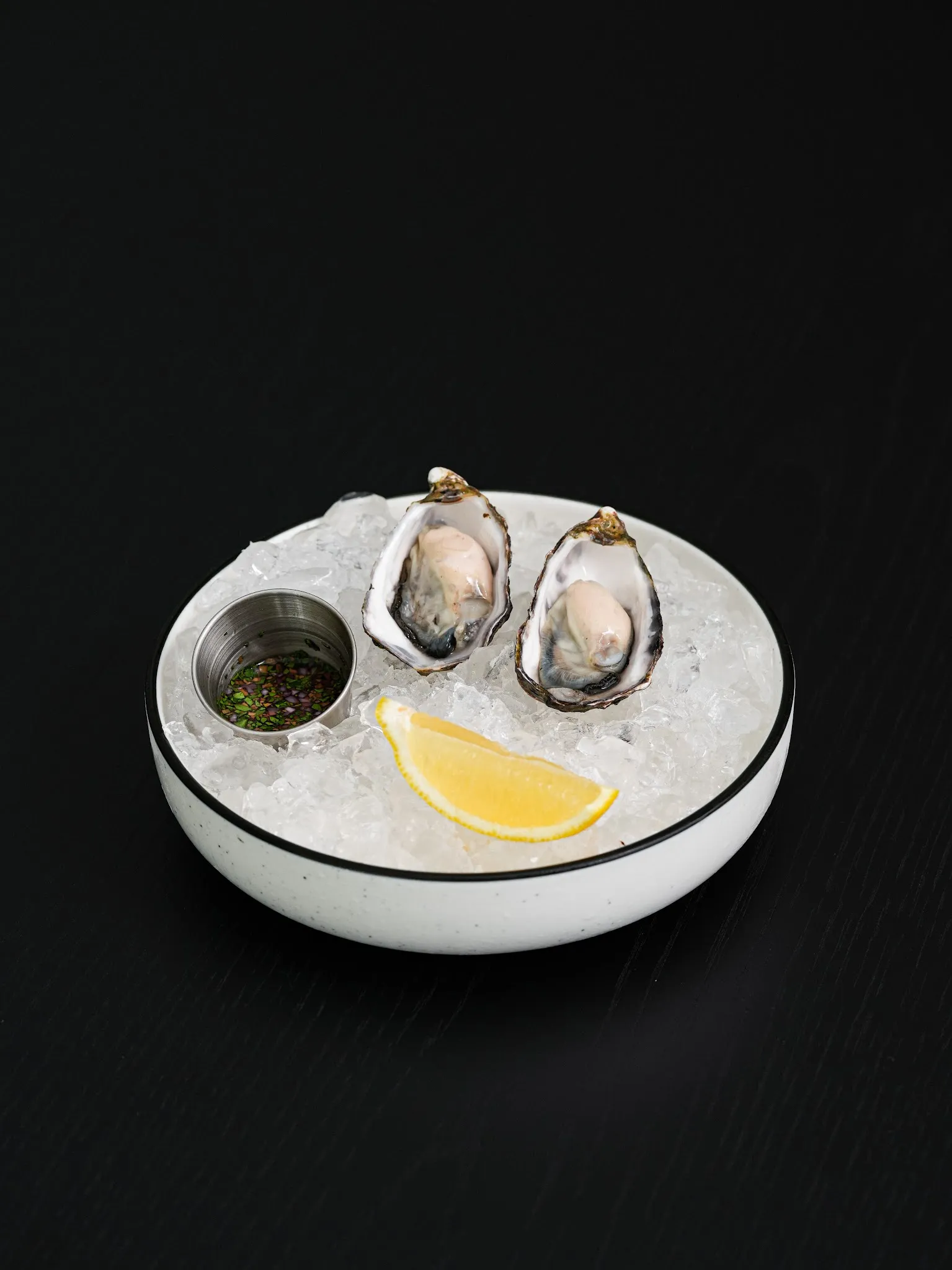 Two fresh oysters on crushed ice with lemon wedge and mignonette in a white bowl