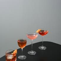 Three cocktails in elegant glassware arranged on a dark surface, blush coupe, amber stem glass, and tumbler with orange