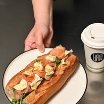 Focaccia sandwich being placed on a white plate next to a Una Una branded takeaway coffee cup