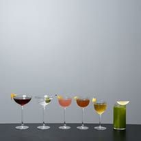 Row of six Una Una cocktails in varied glassware on a light grey surface, spanning the full colour spectrum